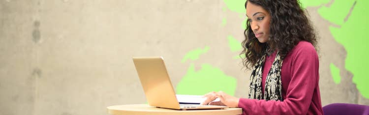 A woman with dark, curly hair wearing a maroon sweater and patterned scarf works on a laptop at a table.