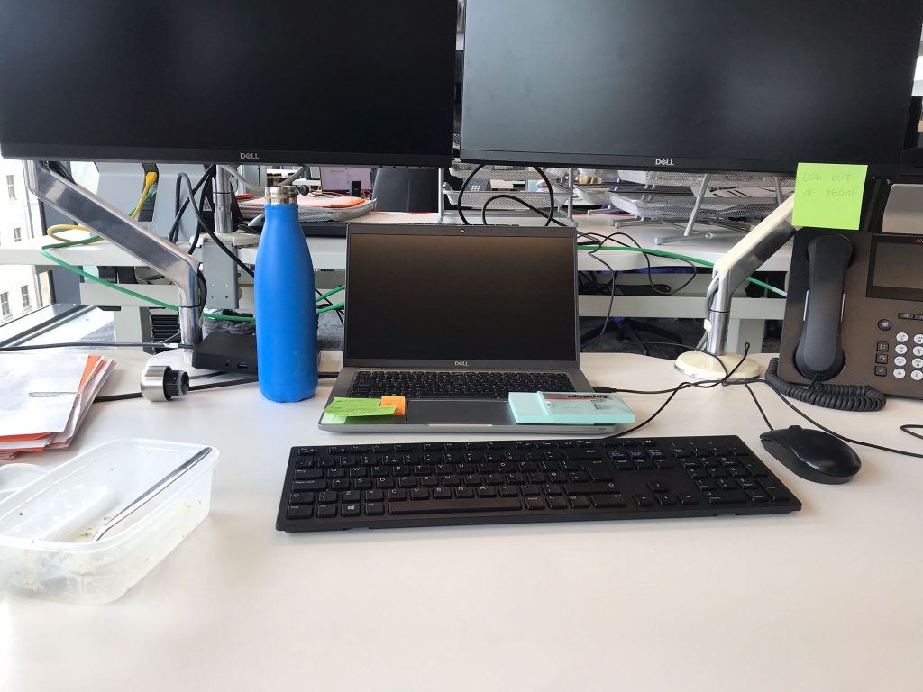 Two desktop screens and a keyboard on a desk with an empty tupperware box and blue water bottle to the right.