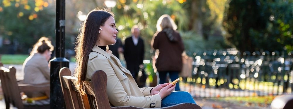 A young professional woman with headphones on is sat outside on a bench.
